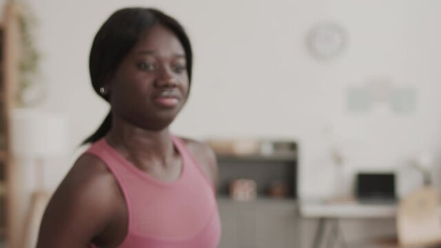 Lockdown of young African-American sportswoman wearing pink sleeveless t-shirt pumping arms muscles using dumbbells while standing in living room