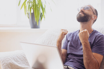 Excited young business man student doing winner gesture, sitting with laptop on sofa at home, in...