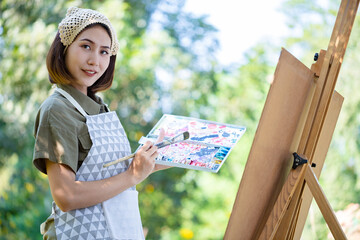 Asian beautiful woman in a summer field with drawing brush and colorful paints on paper artboards in the garden. Left-handed
