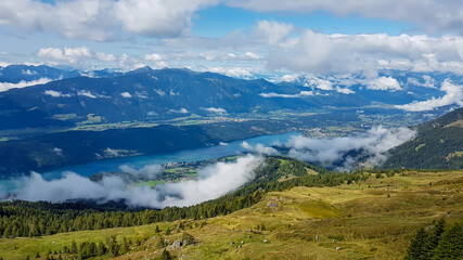 A panoramic view on the Millstaettersee from Granattor in Austrian Alps. The distant lake is surrounded by high mountains. There are few clouds above the lake and the valley. Endless mountain chains