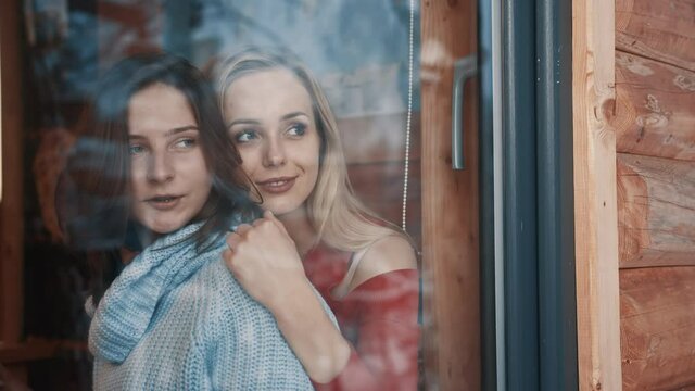 Two women couple on winter vacation. Huggin and looking through the window on snow covered neighborhood. High quality 4k footage