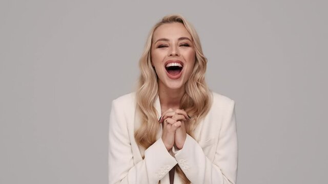 A surprised emotional young blonde businesswoman is looking to the camera and clapping her hands standing isolated over gray background in studio