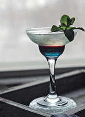 multicolored layered transparent alcohol cocktail in wineglass with mint leaf front view in back light on window background closeup. Selelctive focus