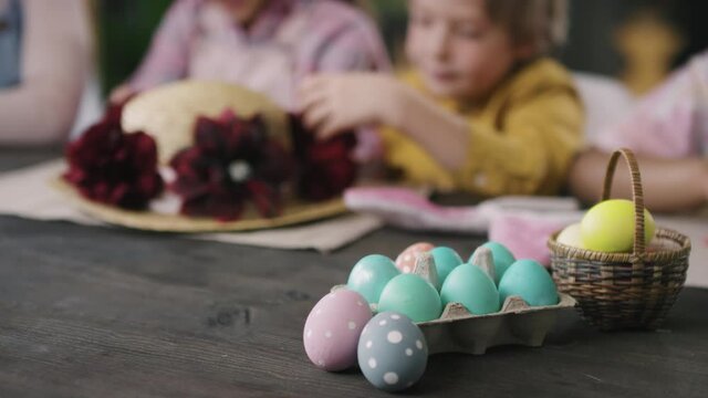 Close up shot of dyed Easter eggs in carton and basket are on wooden table. Children decorating straw hat with flowers in background