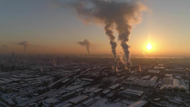 Aerial view of huge factory with tall pipes at sunset. Smoke is coming from the chimneys. Calm weather, smoke accumulates in the air. Winter evening, the yellow sun goes down. Snow on the roofs of bui