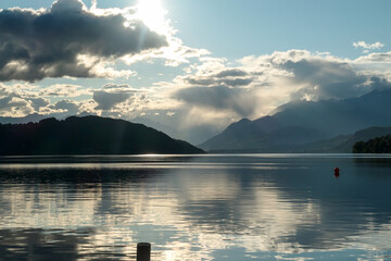A sunset by Millstaetter lake in Austria. The lake is surrounded by high Alps. Calm surface of the lake reflecting the sunbeams. The sun sets behind the mountains. A bit of overcast. Natural beauty
