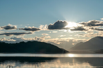 A sunset by Millstaetter lake in Austria. The lake is surrounded by high Alps. Calm surface of the lake reflecting the sunbeams. The sun sets behind the mountains. A bit of overcast. Natural beauty