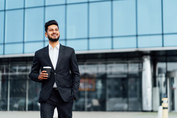 Successful and happy Indian man, businessman, smiling happily going to a business meeting, behind him a modern office building