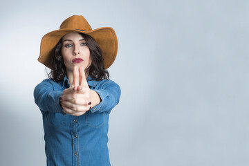 Donna bianca con i capelli neri vestita con una camicia in jeans e un cappello marrone, simula la sparatoria con e dita  , isolata su sfondo bianco