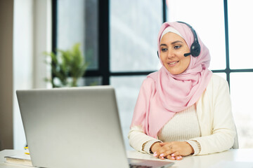 A middle-eastern woman wearing national hijab using a laptop and a wireless headset for connection and support customers in the office. Concept of diverse call center employees, multiracial team