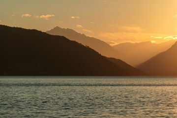 A sunset by Millstaetter lake in Austria. The lake is surrounded by high Alps. Calm surface of the lake reflecting the sunbeams. The sun sets behind the mountains. A bit of overcast. Natural beauty