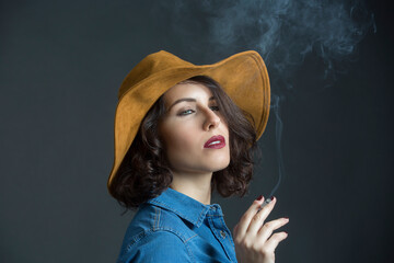 Donna mora con camicia in jeans e cappello da cowboy, fuma una sigaretta con espressione seria , isolata su sfondo grigio