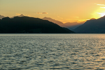 A sunset by Millstaetter lake in Austria. The lake is surrounded by high Alps. Calm surface of the lake reflecting the sunbeams. The sun sets behind the mountains. A bit of overcast. Natural beauty