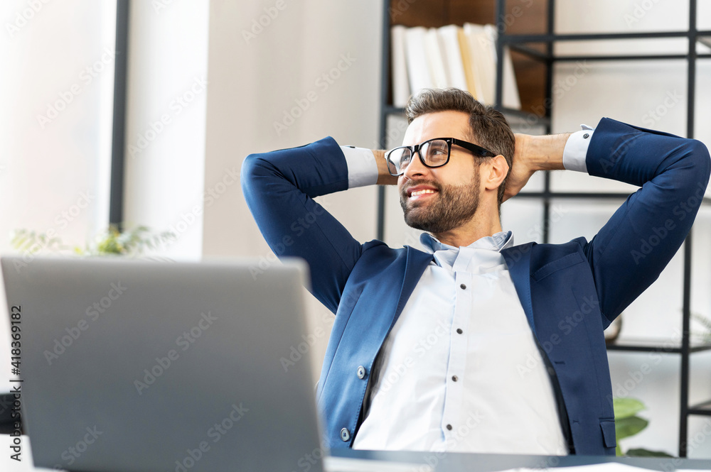 Wall mural Smiling man with glasses sitting at the laptop, holding hands behind head, relaxing, looking at good weather outside the window, works in a comfortable office, did the job productively and got praise