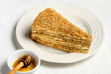 Russian honey cake slice on white plate on off white textile tablecloth. Traditional 8 layer delicious dessert from former Soviet Union. Honey in small jar with wooden stick.