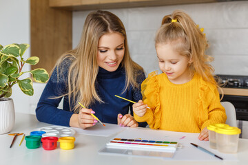 Mother and daughter painting at home. Cute little kid in yellow sweater having fun with parent and paints. Concept of early childhood education, hobby, talent, preschool leisure and parenting