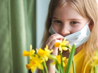 Coronavirus is over. Little girl sitting on the windowsill and removing face medical mask, sniffing yellow daffodil flowers. Take off the mask. Quarantine is over. Hello spring