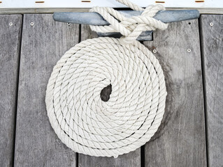 White rope coiled on a weathered wooden dock and tied to a metal cleat.  Closeup.