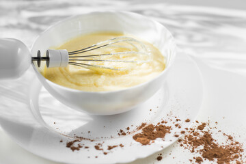 Preparation of cake cream. Cream whipped with mixer in a plate on a white background. Сocoa scattered on a table. Light rays fall on the table