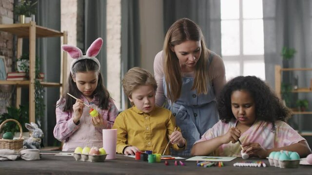 Medium shot of cheerful female teacher talking and checking work of cute children making Easter cards and decorating eggs in arts and crafts class