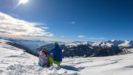 A couple sitting on the snow on top of Katschberg in Austria. They are happy and joyful. Panoramic view on the surrounding mountains. Winter wonderland. Bright and sunny winter day. Love and happiness