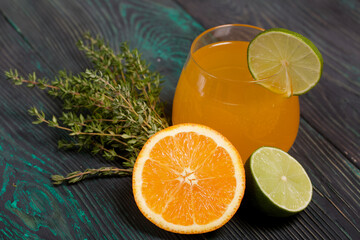 A glass of alcohol. Decorated with a lime wedge. Nearby halves of lime, orange and thyme.