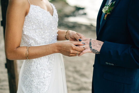 Bride putting on groom's wedding ring on lakeside, mid section