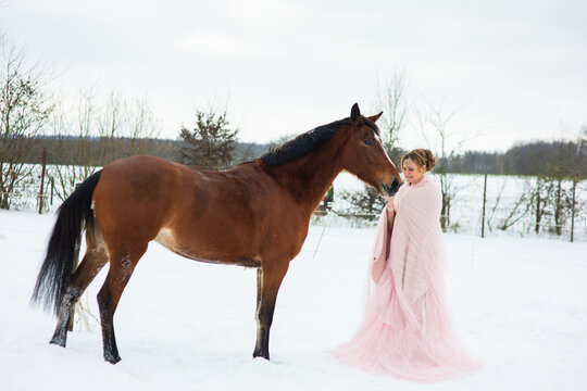 Eine h&uuml;bsche Frau, sieht aus wie Aschenputtel steht in Schnee mit Rosa  T&uuml;ll Kleid und streichelt ihr Pferd