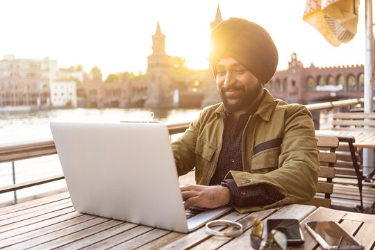 Indian man using laptop in cafe by river, Berlin, Germany - Powered by Adobe