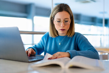 Female higher education student using laptop and studying book in university cafe