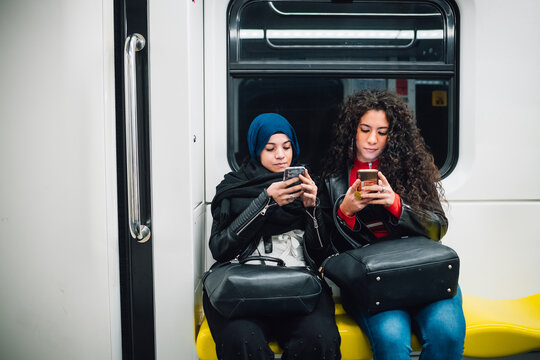 Young woman in hijab and friend sitting on subway train looking at smartphones