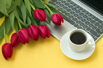 laptop, a bouquet of red tulips, a cup of coffee on a yellow background. a gift to a colleague on March 8.