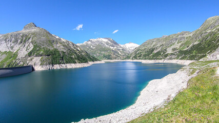 Dam in Austrian Alps. The artificial lake stretches over a vast territory, shining with navy blue color. The dam is surrounded by high mountains. In the back there is a glacier. Controlling the nature