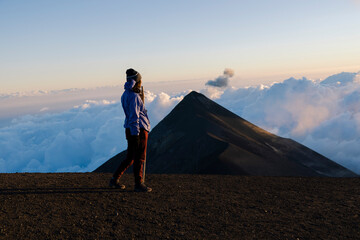Young woman on top of the Acatenango volcano in Guatemala with the Volcano of Fuego in the background - woman watching the sunset in the mountain range