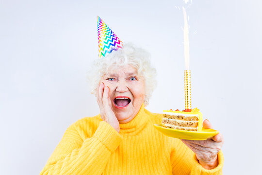 Older Woman With A Gift Wear Yellow Sweater And Horn Cap On A White Background Holding Plate With Cake With Fireworks