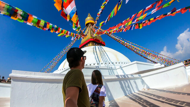 A couple walking along the prayer flags hanging from the top of boudhanath Temple in Kathmandu, Nepal. The flags have 'om mani padme hum' mantra written on them. The round temple is painted white.