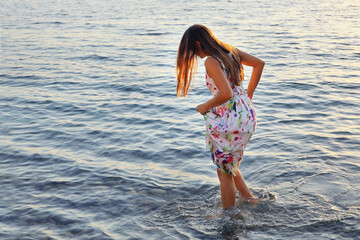 Young woman in dress walks slowly barefoot on water at sunset on summer day