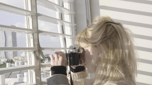 Woman taking pictures from window. Young beautiful lady peeking out of curtains and looking down to the street. Beautiful sun ray through shutters on blond ladies face. shooting pics from old camera