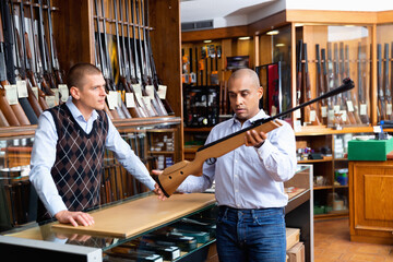 Hispanic man examining hunting break-barrel air rifle in gun shop, consulting with salesman
