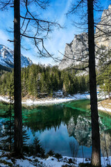 Obraz premium An idyllic view on Alpine Green Lake in Austria, seen from a small hill around the lake, through the trees. Powder snow covering the mountains and ground. Emerald color of water. Winter Wonderland