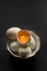 Opened egg with visible yolk on top of other eggs in bowl on black background waiting for easter