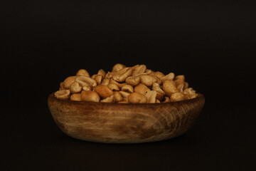 Close up of pile of salted, roasted peanuts in a wooden bowl on black background
