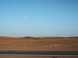 Desert asphalt route and Moon over desert landscape at early evening. Blue, brown, black, white...