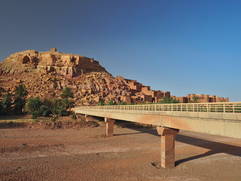 View of Aint Ben Haddu old city in Morocco, UNESCO list. Bridge over periodic river. Red brick, green palms, blue sky
