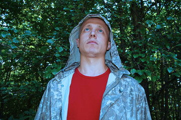 Portrait of a young man in a camouflage jacket looks up against the background of green foliage in...