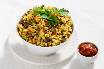 Plov, pilaf or pilau, rice dish from Central Asia made with golden rice, carrot and browned lamb meet. The portion is served in ceramic bowl on off white textile tablecloth. Ajika sauce on the side.