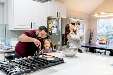 Dad making pancakes with toddler daughter, mom holding baby