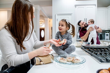 Mom and daughter baking cinnamon rolls in kitchen while dad plays with baby in the background