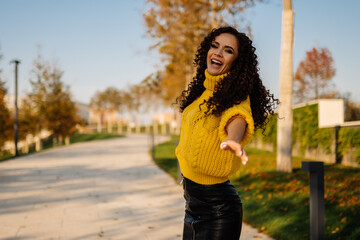 On the track in the park sings actively putting forward one hand with a wide open mouth curly brunette in a bright sweater and leather skirt. High quality photo