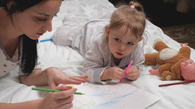 Mother, nanny, teaches child girl to draw. Happy family playing together at home on bed. Mom helps her daughter learn to draw on paper by coloring with multi-colored pencils and felt-tip pens.